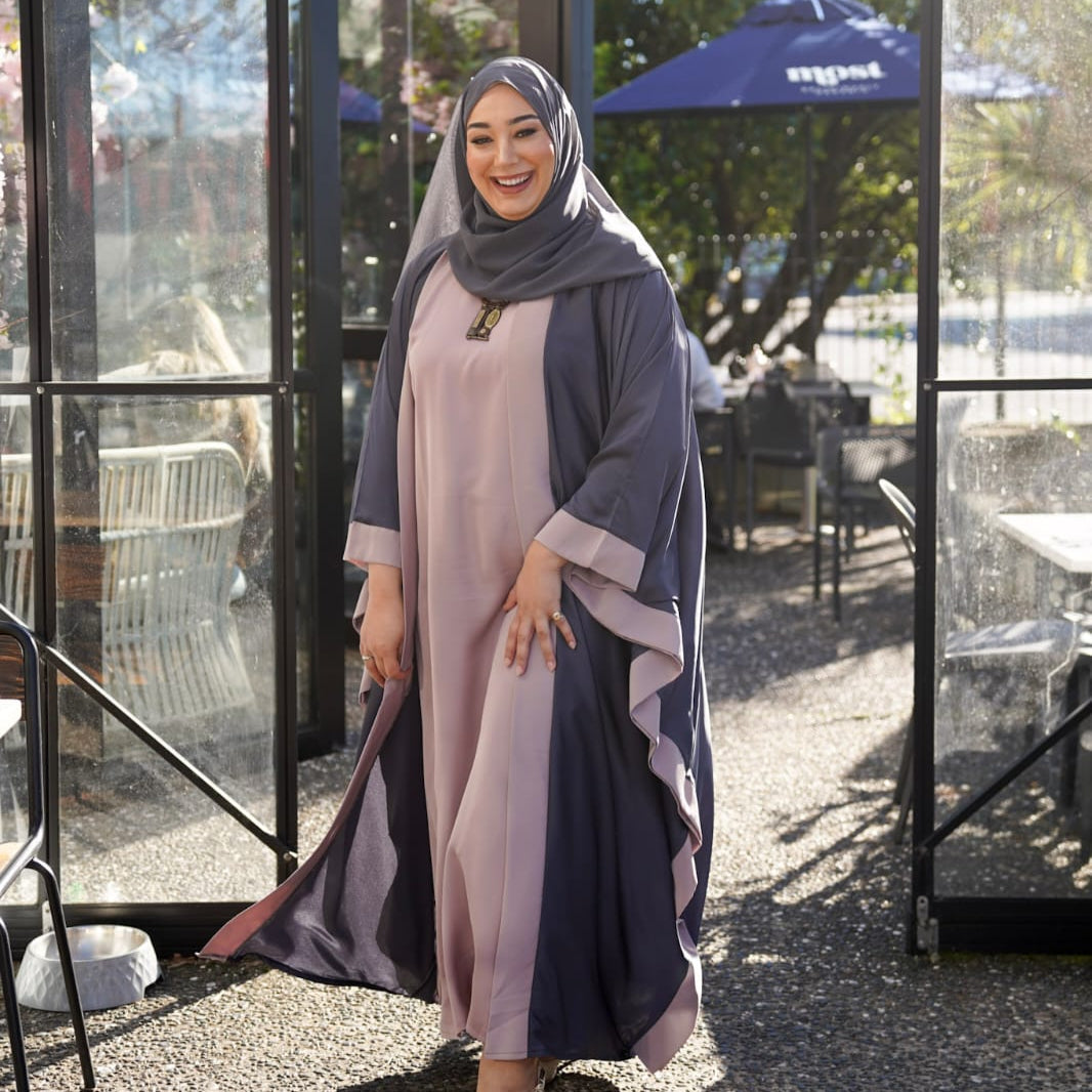 Muslim woman wearing a modest fashion outfit featuring a dark grey abaya with light trim, pink inner dress, matching scarf hijab, and high heels — showcasing modest fashion in an outdoor Auckland café setting, Fauz Closet New Zealand