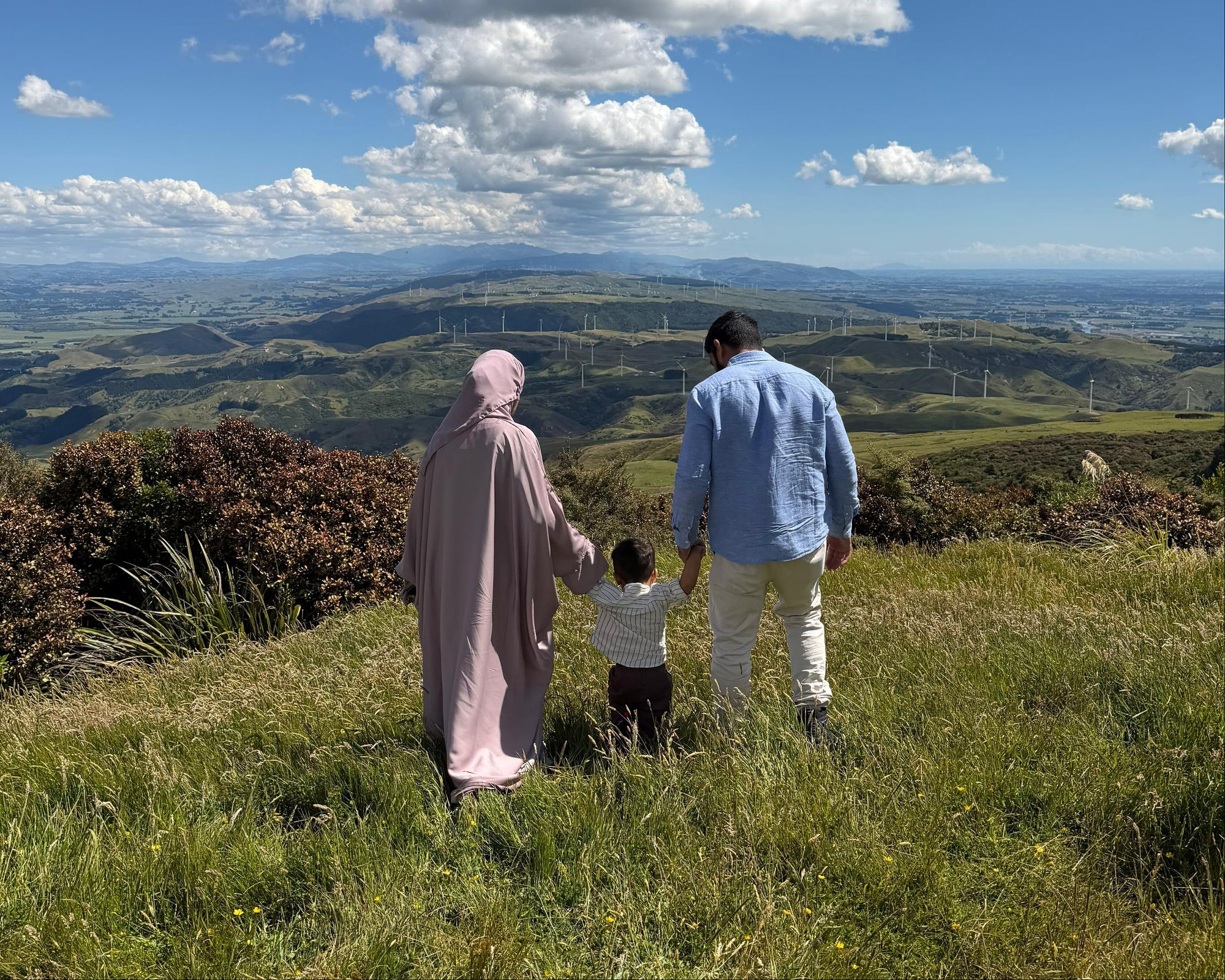 A small family of three walking on a grassy hill with a scenic view of rolling hills and blue sky.